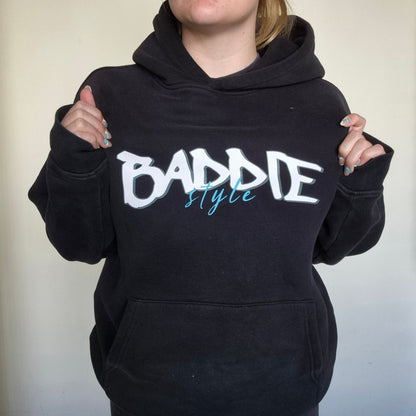 Person wearing a black hoodie with 'BADDIE STYLE' text on a plain background
