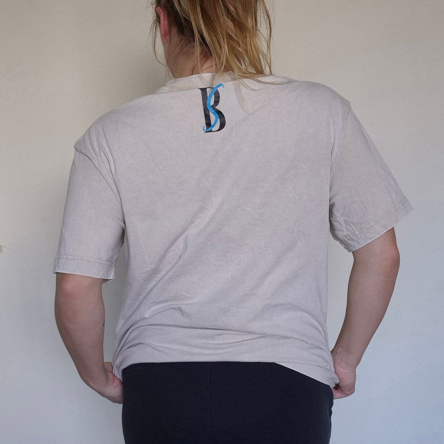 Person wearing a light gray t-shirt with a logo on the back, standing against a plain white wall.