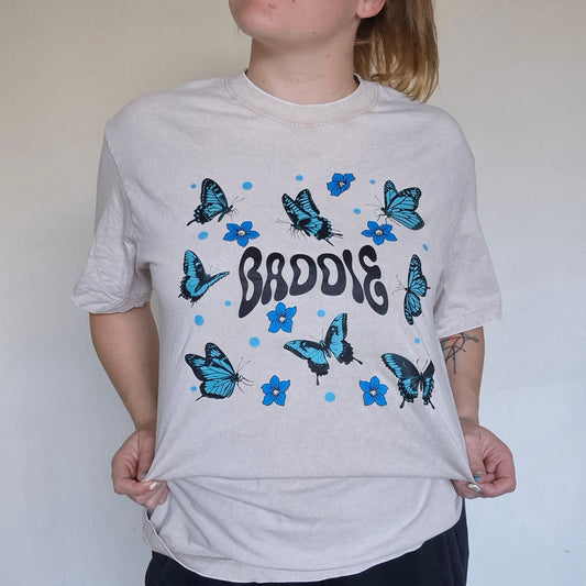 Person wearing a white t-shirt with blue butterfly designs and text, standing against a plain wall.