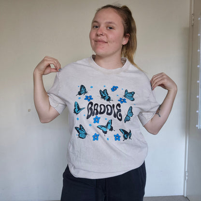 Person wearing a white t-shirt with blue butterfly designs and text, standing against a plain wall.