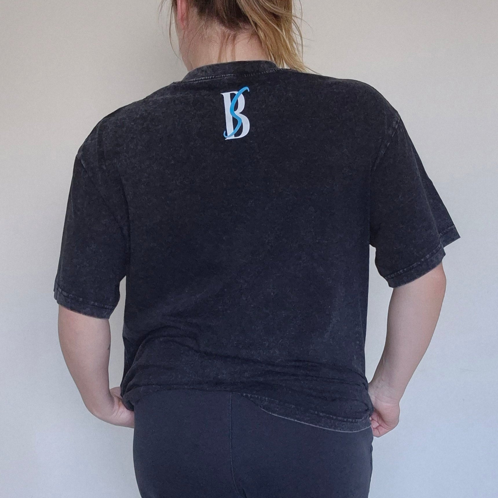 Person wearing a black t-shirt with a logo on the back, standing against a plain white wall.