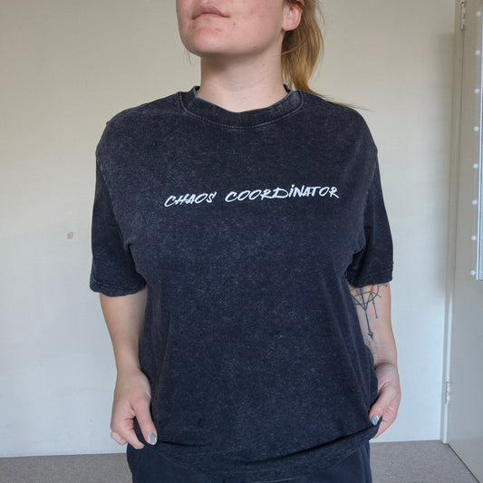 Person wearing a black t-shirt with 'CHAOS COORDINATOR' text in a plain room.