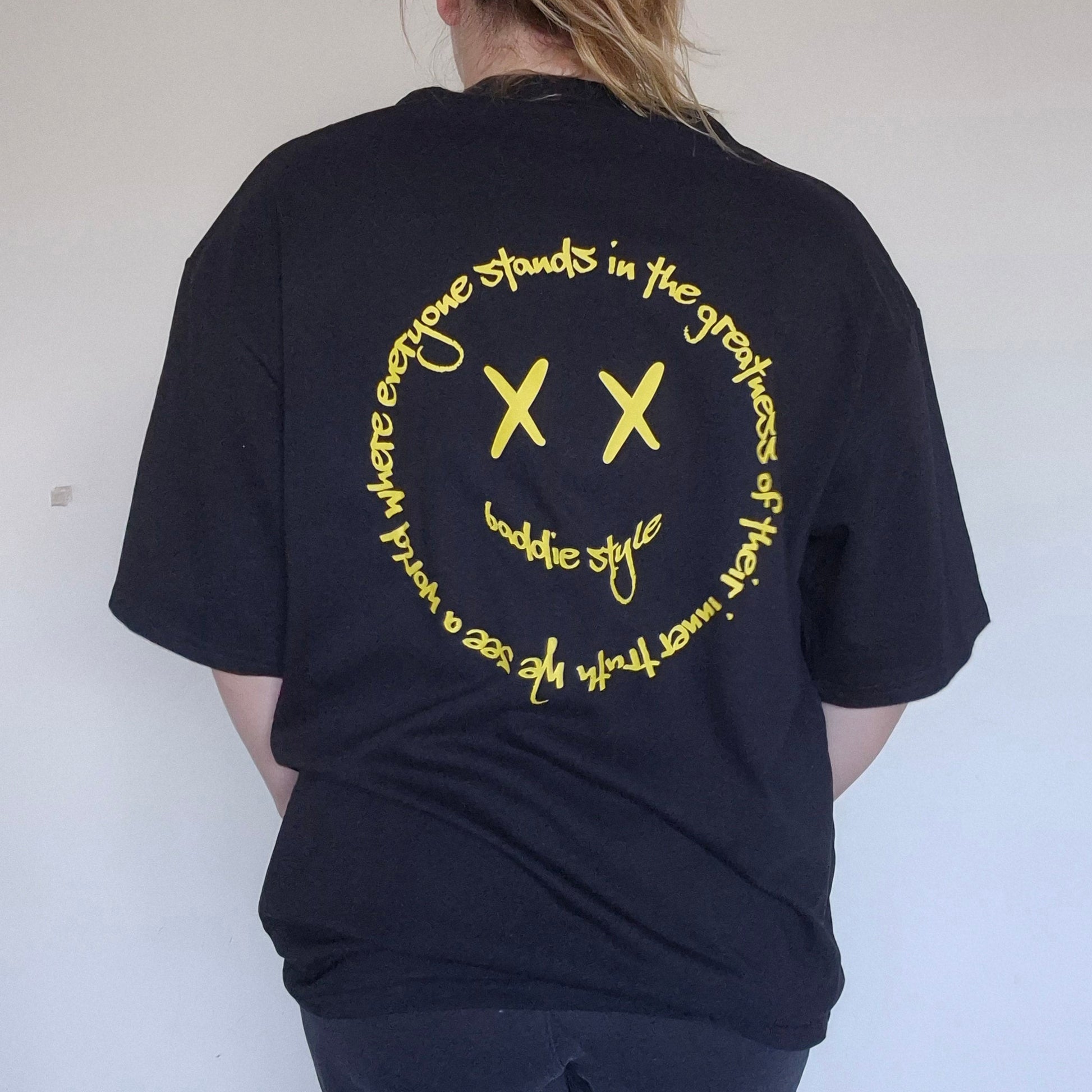 Person wearing a black t-shirt with yellow text and symbols on the back, standing against a plain white wall.