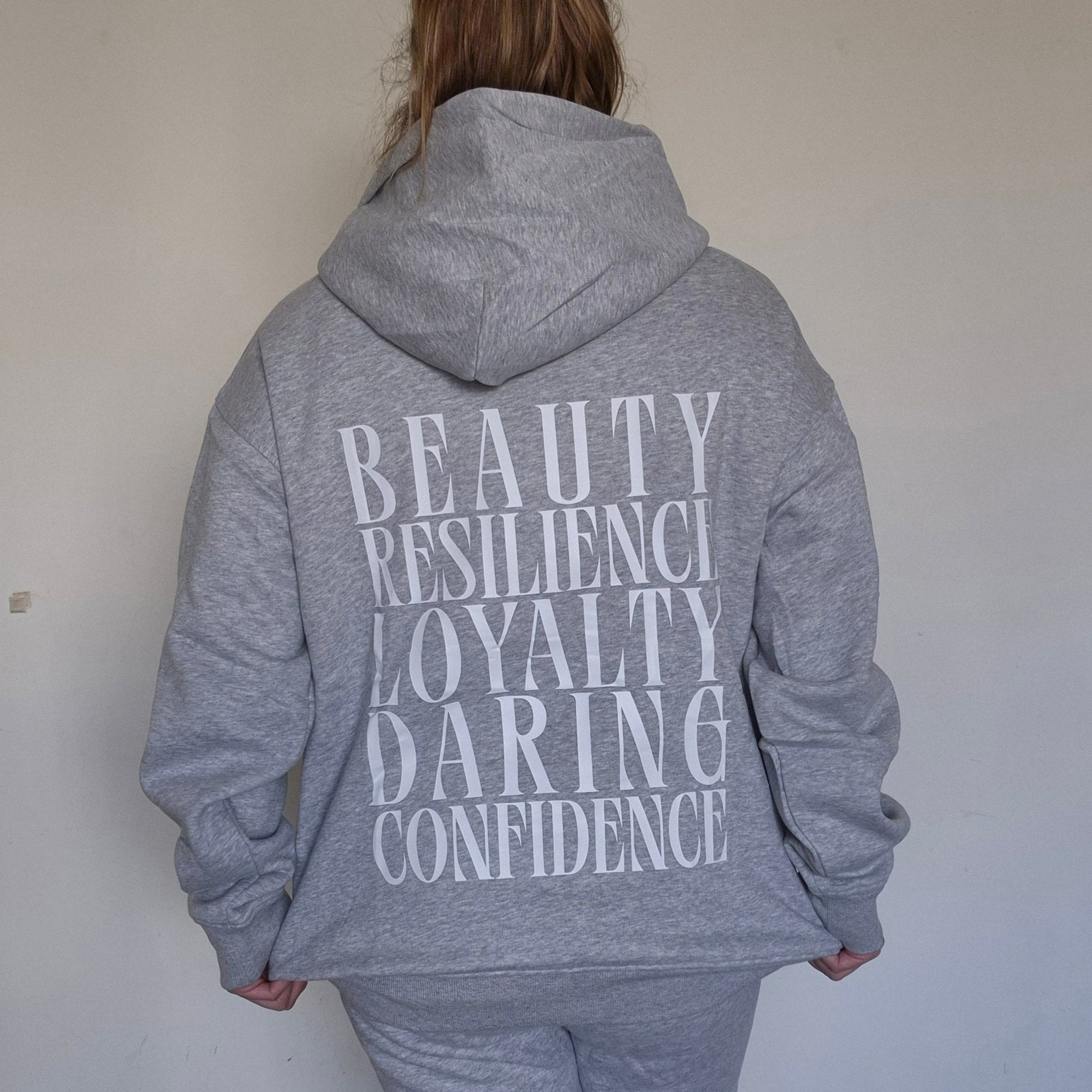 Person wearing a gray hoodie with motivational text on the back against a plain wall.