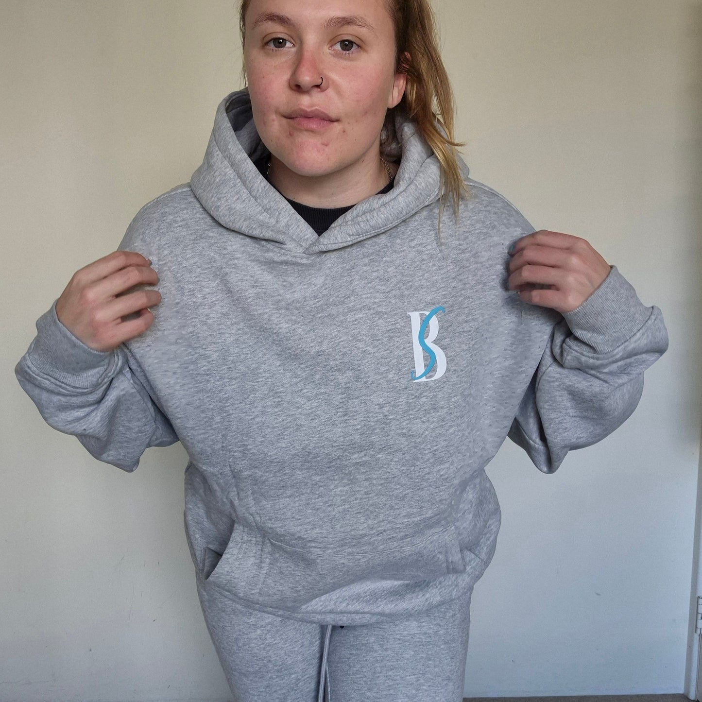 Person wearing a gray hoodie with a logo, standing against a plain background